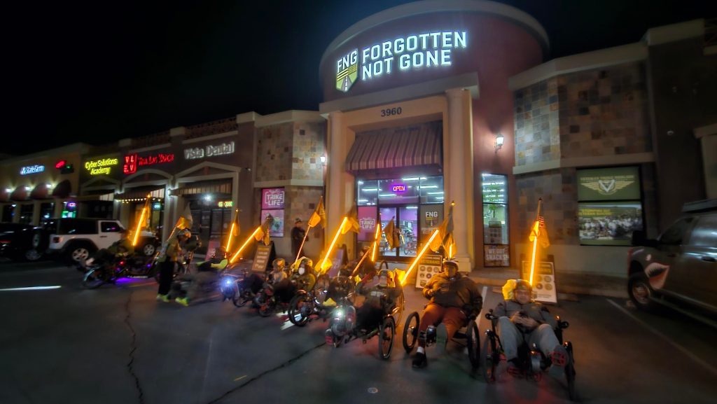Veterans on trikes with glowing orange flags in a parking lot at night in front of the Forgotten Not Gone Headquarters and Trike Life Shop.