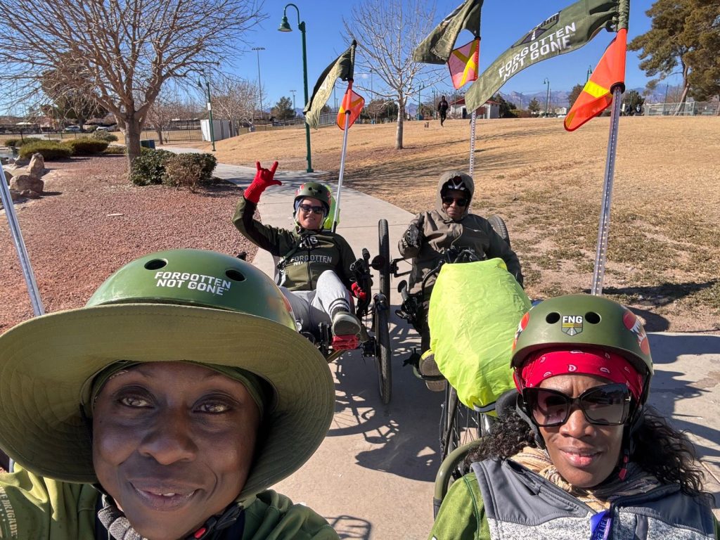 Four women veterans riding adaptive trikes on a sunny day during the Women Warrior Ride, wearing Forgotten Not Gone helmets and gear. The image captures camaraderie and smiles, with FNG flags prominently displayed in the background against a clear blue sky.